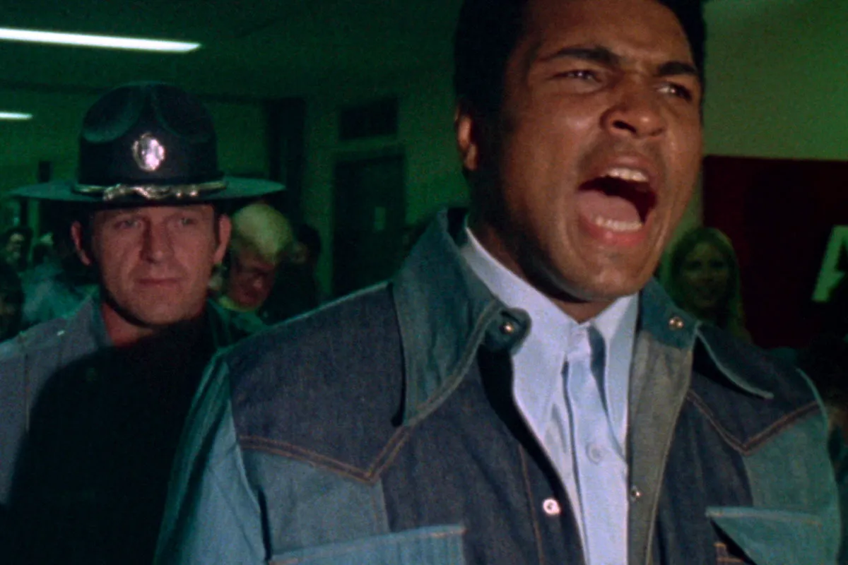 Boxer Muhammad Ali is a Black man in a blue shirt with a denim jacket . He is being arrested by a white policeman.From Leon Gast’s 'When We Were Kings.' Courtesy of The Criterion Collection.