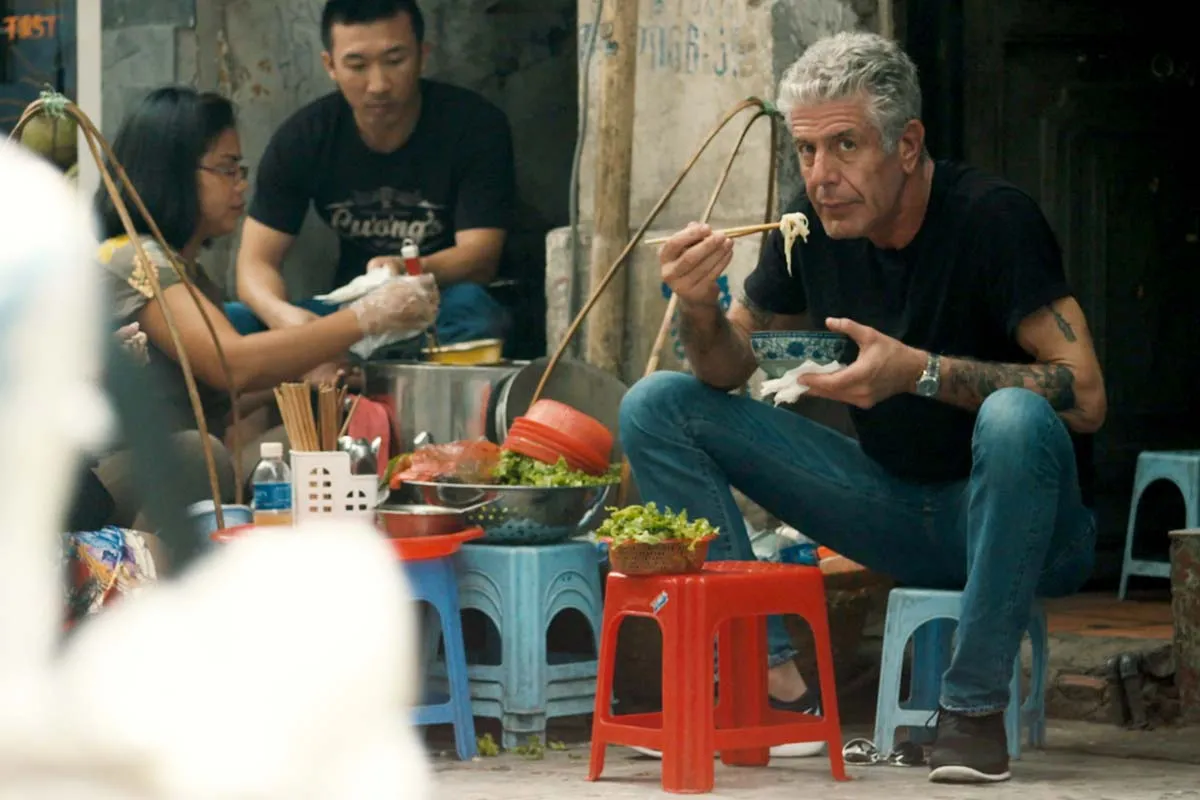 Anthony Bourdain, a white man with white hair, a black short and tattoos on his arms, sits on a sidewalk, eating a bowl of noodles. From  Morgan Neville's  'Roadrunner.' Courtesy of CNN / Focus Features