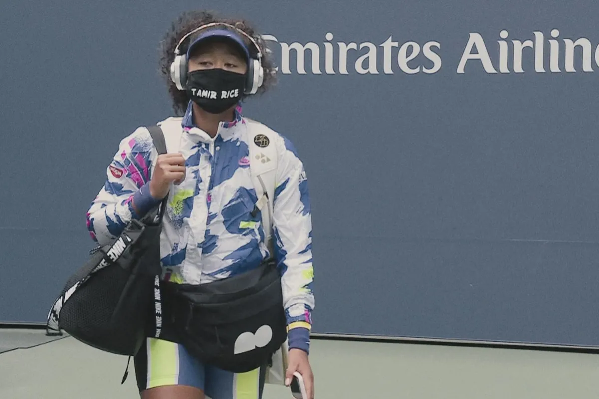 Tennis player Naomi Osaka, an Afro-Japanese female tennis player on a tennis court wearing a multi-colored jacket, and a mask that says “Tamir Rice.” Still from Garrett Bradley’s Netflix series, ‘Naomi Osaka’. Courtesy of Netflix
