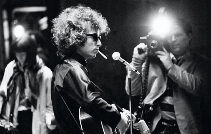 Bob Dylan, a white man with curly hair, performs on stage with a photographer snapping a photo. A still from 'Don't Look Back'. Courtesy of Pennebaker Hegedus Films
