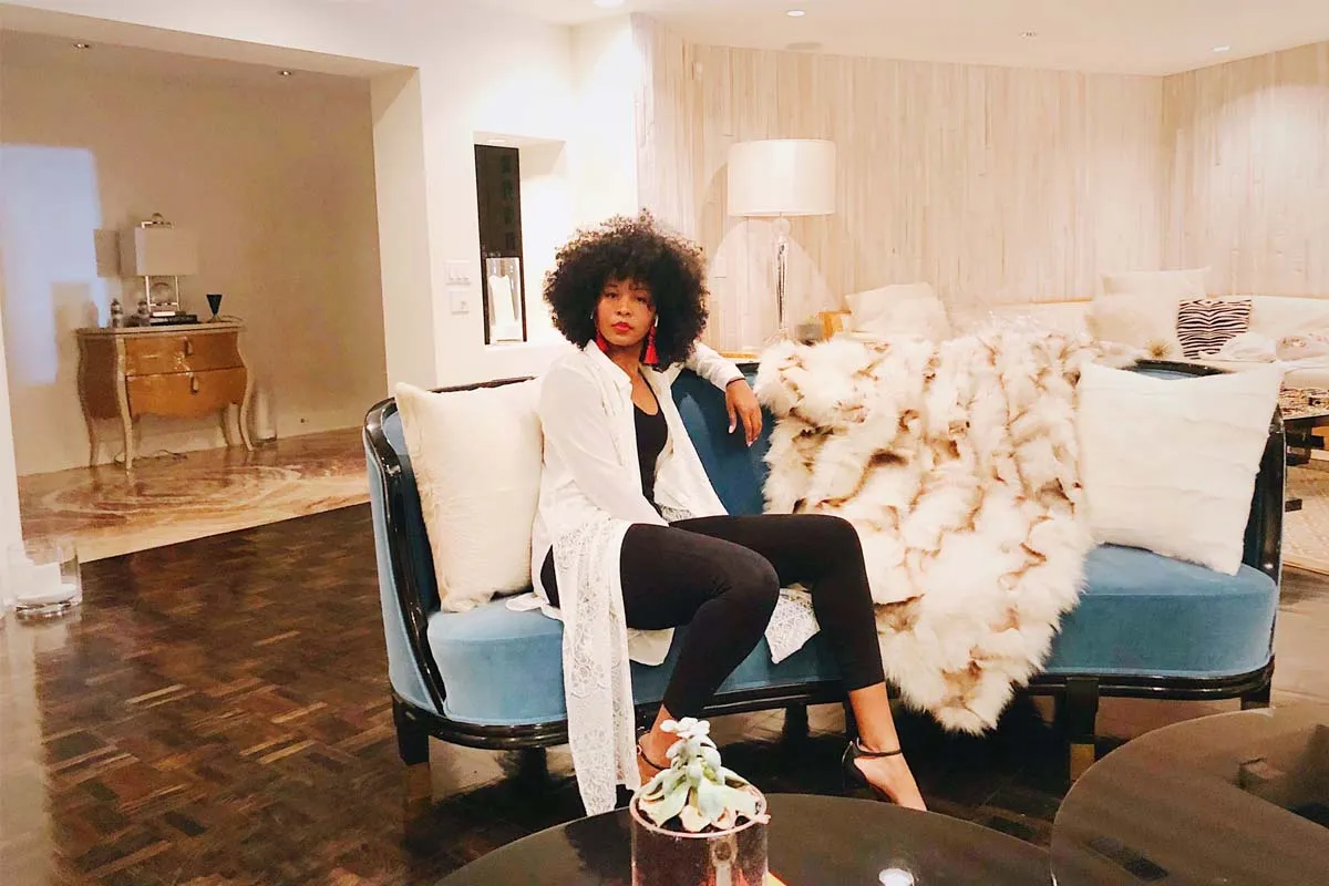  A full body shot of a Black woman in a black bodysuit and sheer white cover-up with black high heels and red dangling earrings, sitting on a couch with an afro.