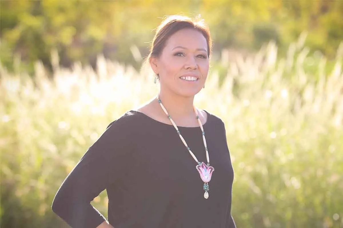 Headshot of a Mohawk woman with medium skin tone, brown hair worn up, wearing a black shirt and a pink beaded floral necklace 