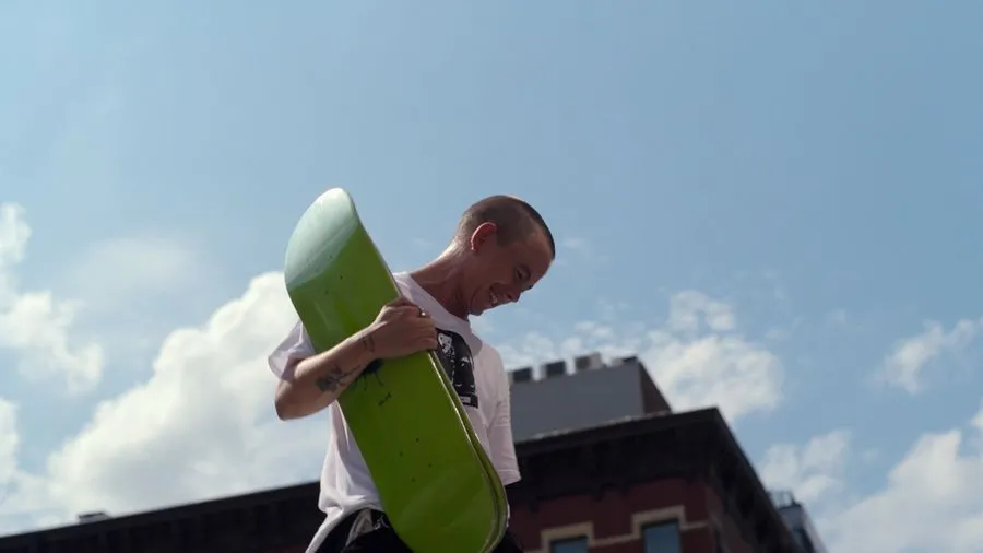 Leo Baker, a white person with buzz-cut hair, smiles toward the ground while holding a neon green skateboard deck. From Nicola Marsh and Giovanni Reda’s  ‘Stay on Board: The Leo Baker Story,. Photo courtesy of Netflix.