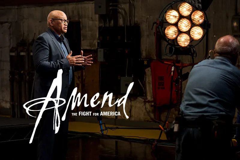 Larry Wilmore, an African American male film director, is wearing a black blazer over a blue shirt, talking to someone off screen on a film set. 