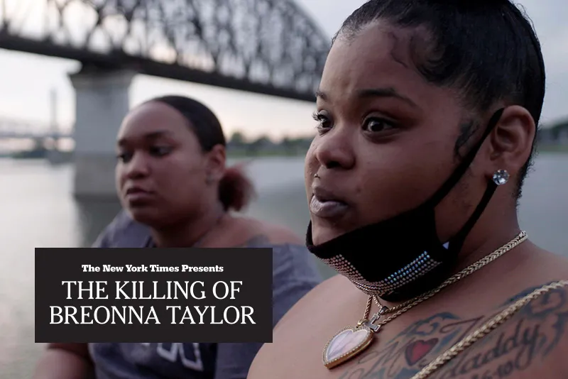 Two adult women with dark skin tone are next to a river with a bridge in the background. 