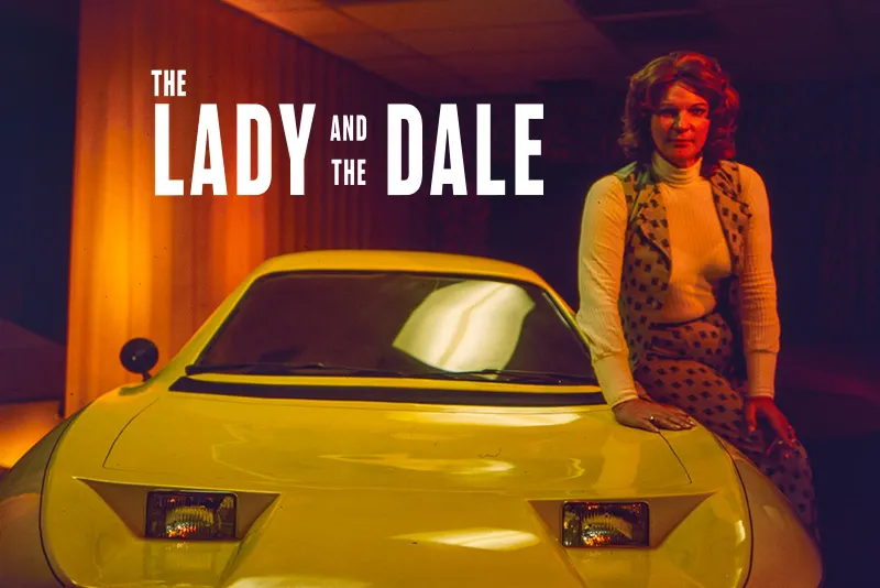 A transgender woman with brown hair to her shoulders is sitting on the side of a yellow car