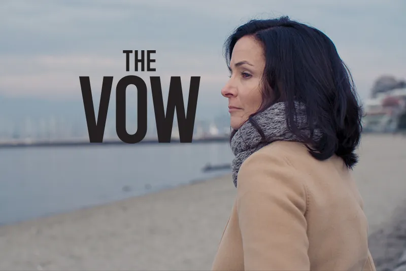 A light-skinned woman, wearing a grey scarf and cream coat, is looking out on a beach