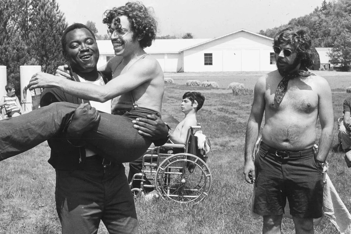 Teenagers with disability at summer camp. A Black man is holding a light-skinned man with curly hair.