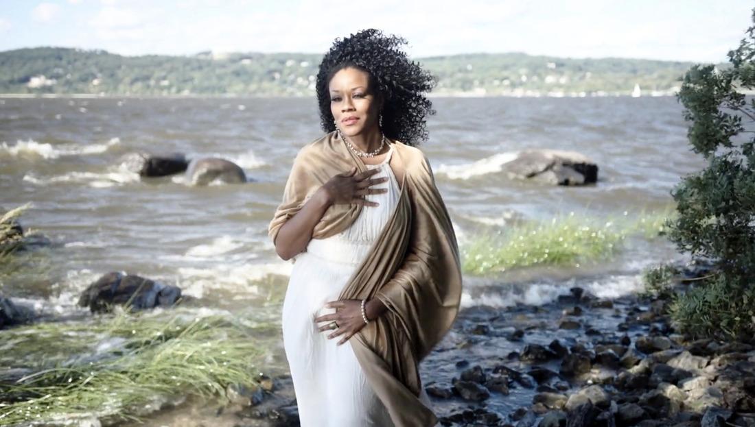 A Black femme in a white dress looks tranquilly toward the camera, as in Botticelli’s Birth of Venus, at the bank of a river.