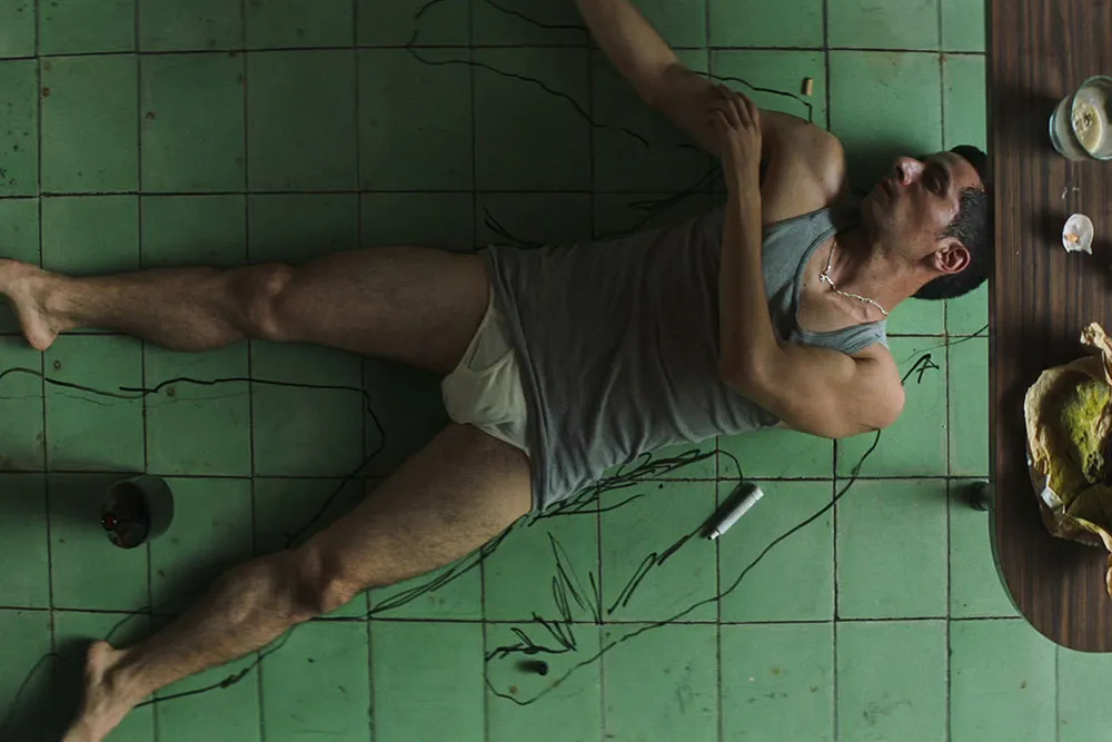A man with brown skin and short hair, wearing a tank top and underwear, traces himself on the floor.