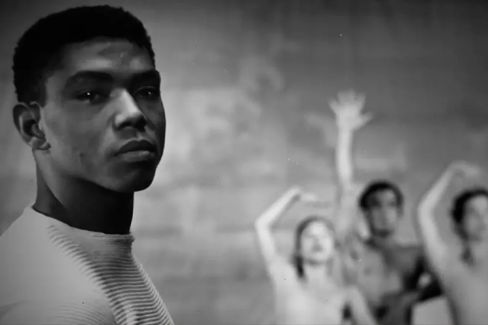 Black and white image of a black man with short dark hair looking into the camera. In the background three dancers strike a pose.