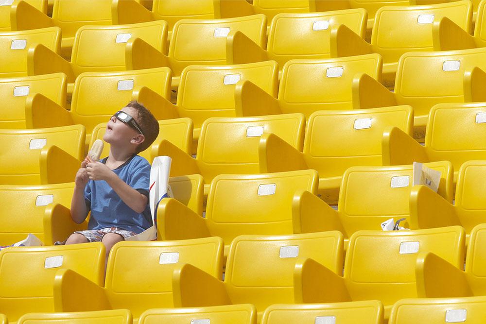 A child with white skin, short hair and wearing a blue shirt and solar viewing glasses. He sits in stadium seating eating a popsicle. 