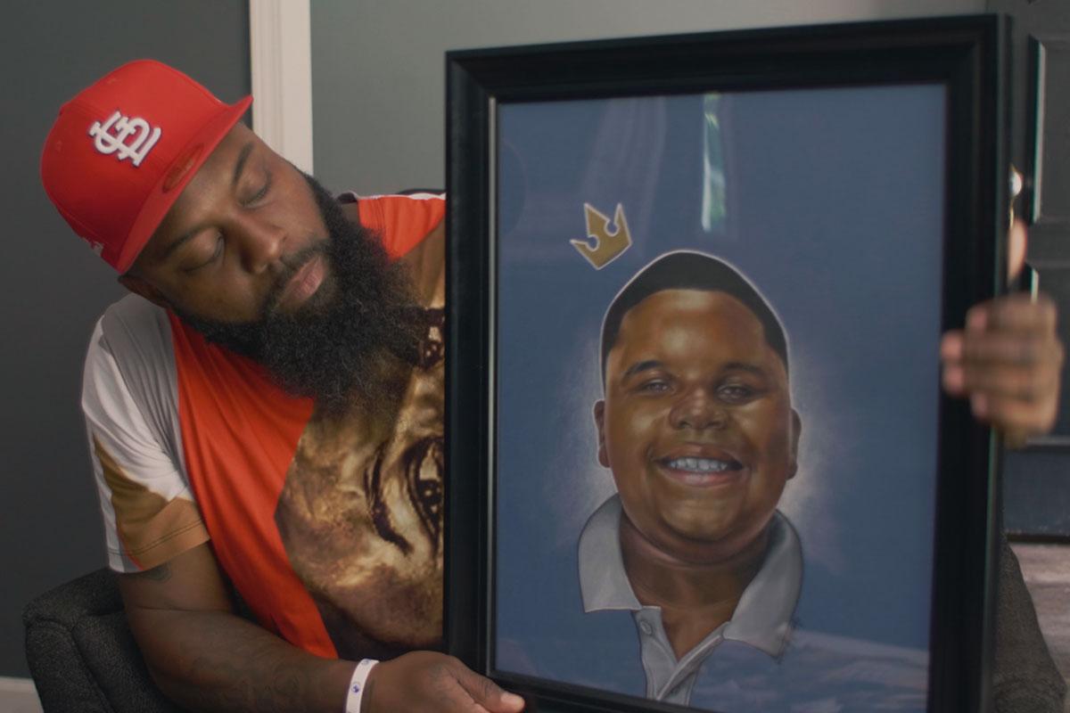 A Black man with a long black beard wearing a red baseball cap and shirt holds a framed painting of a Black boy with a crown on his head.