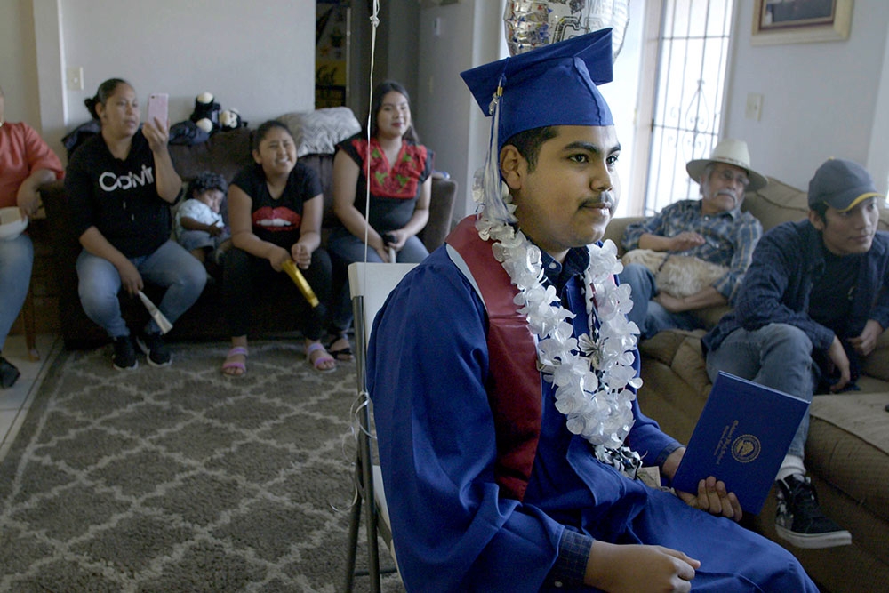 A male student in a blue graduation cap and gown and flower neckless sits among a group of people in a living room.