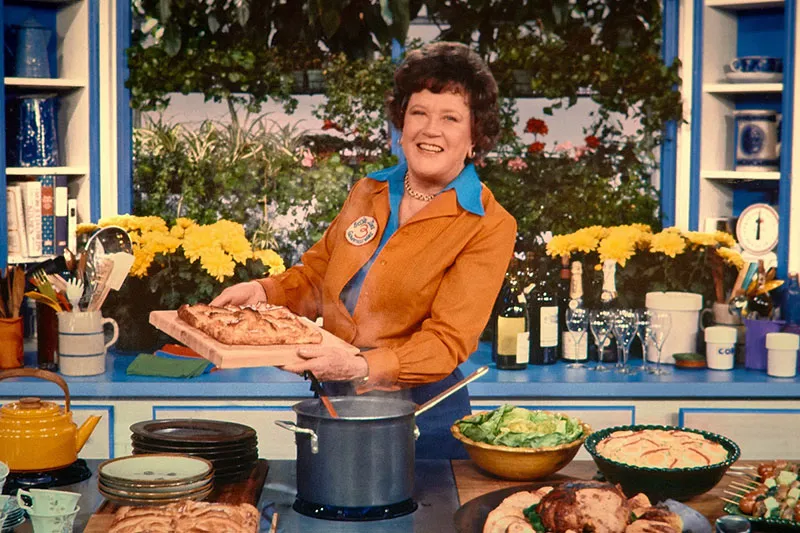 A woman with white skin and curly brown hair. She holds a pan with food on it.