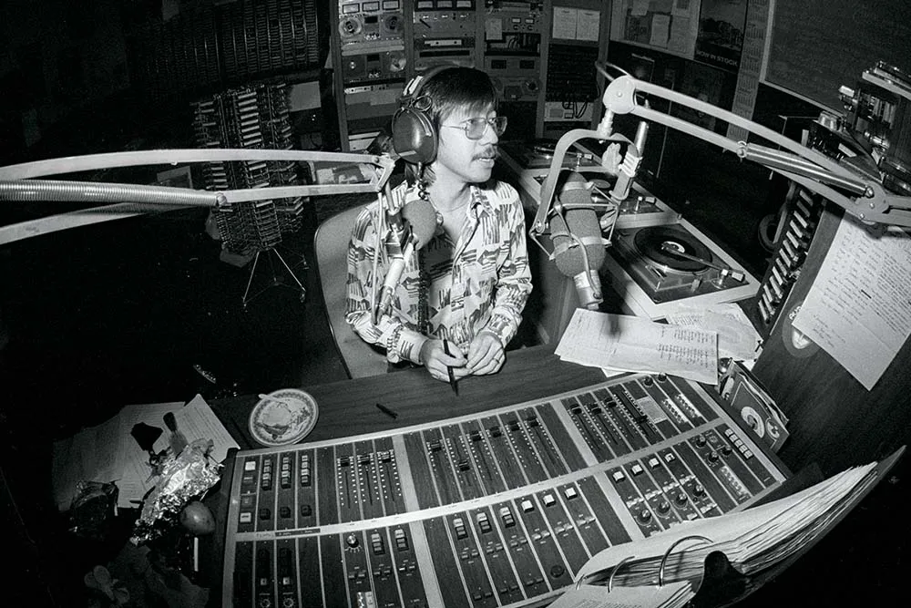 A black and white photo of an asian man in a patterned button up. He sits in a recording studio.