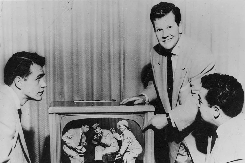 A black and white picture of several men with light skin in suits standing around a TV. 