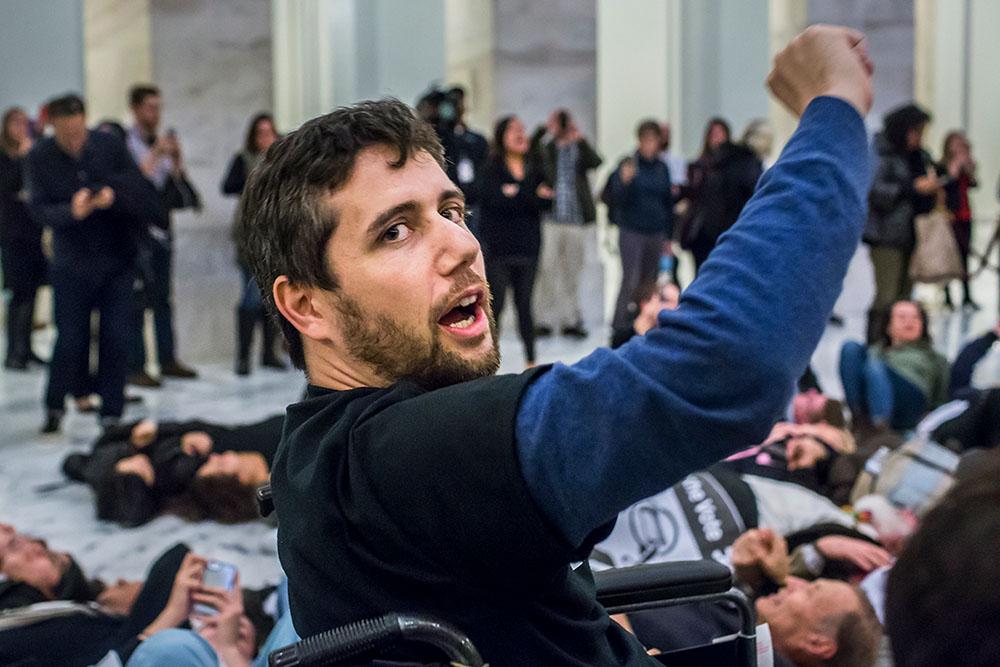 A white man with brown hair and facial hair raises his fist in the air. He is in a wheelchair.