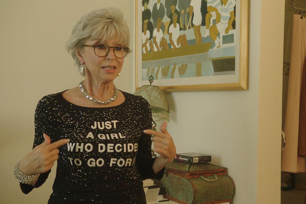 An older white woman with gray hair and black glasses. She is pointing to her shirt which reads "Just A Girl Who Decided To Go For It."