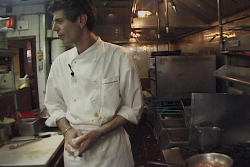 Anthony Bourdain, an older white man with short hair wearing a white chef's coat, stands in a small kitchen.