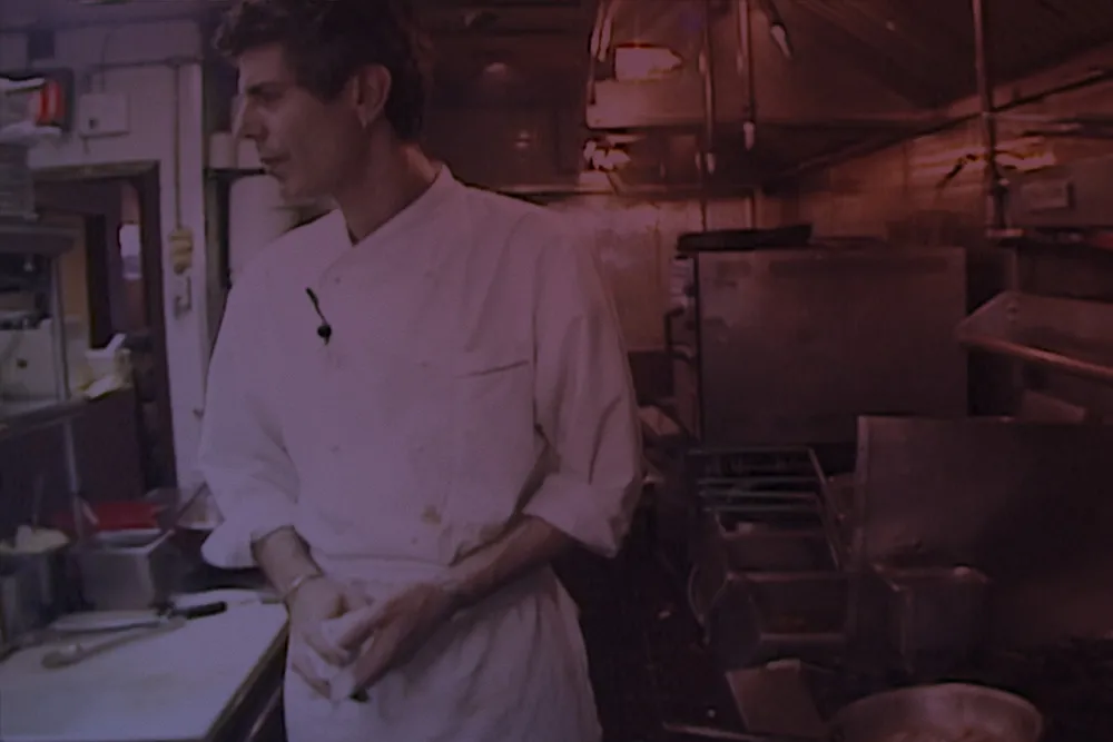 Anthony Bourdain, an older white man with short hair wearing a white chef's coat, stands in a small kitchen.