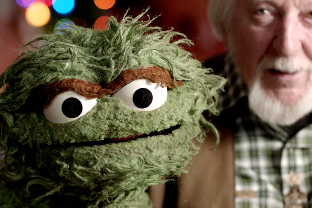 An older white man with gray hair holds up a green, scraggly puppet.