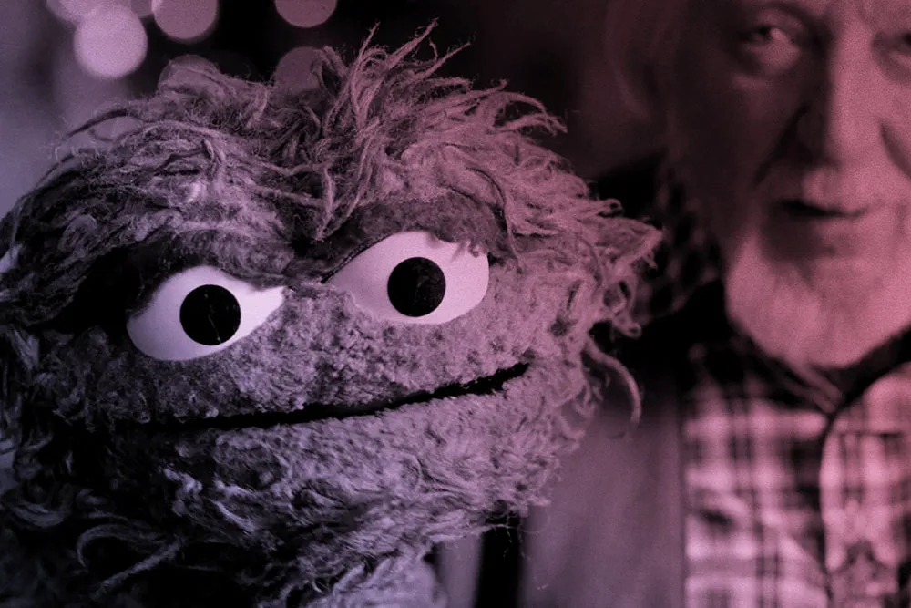 An older white man with gray hair holds up a green, scraggly puppet.