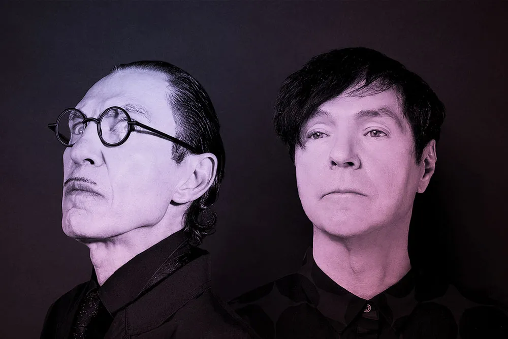 A black and white portrait of two white men both with dark hair. The man on the left has round black glasses and a pencil mustache.