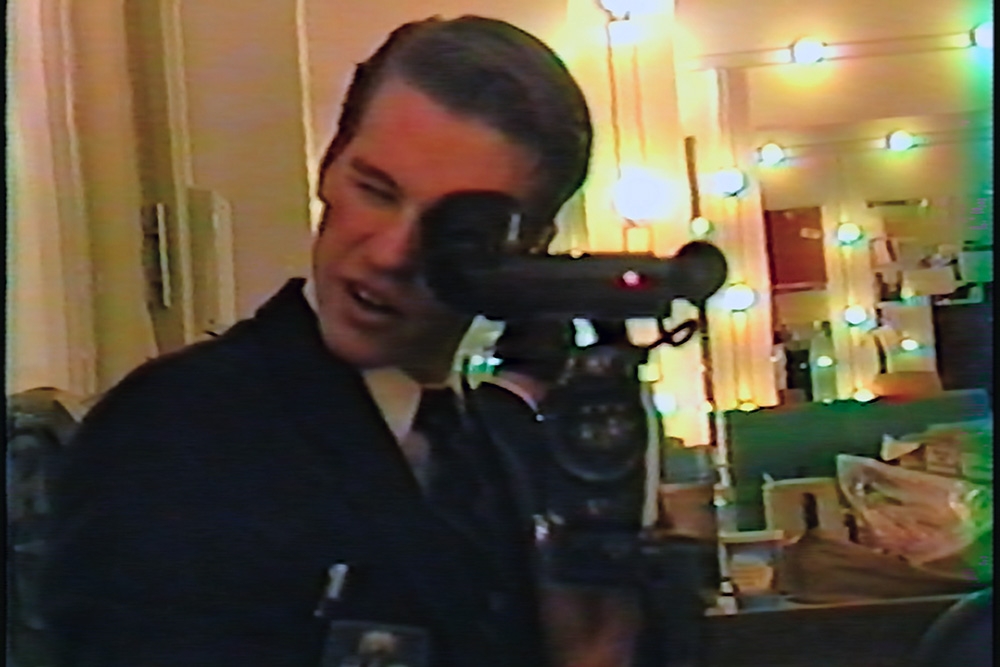 A white man with short brown hair holds a camera up to a mirror.