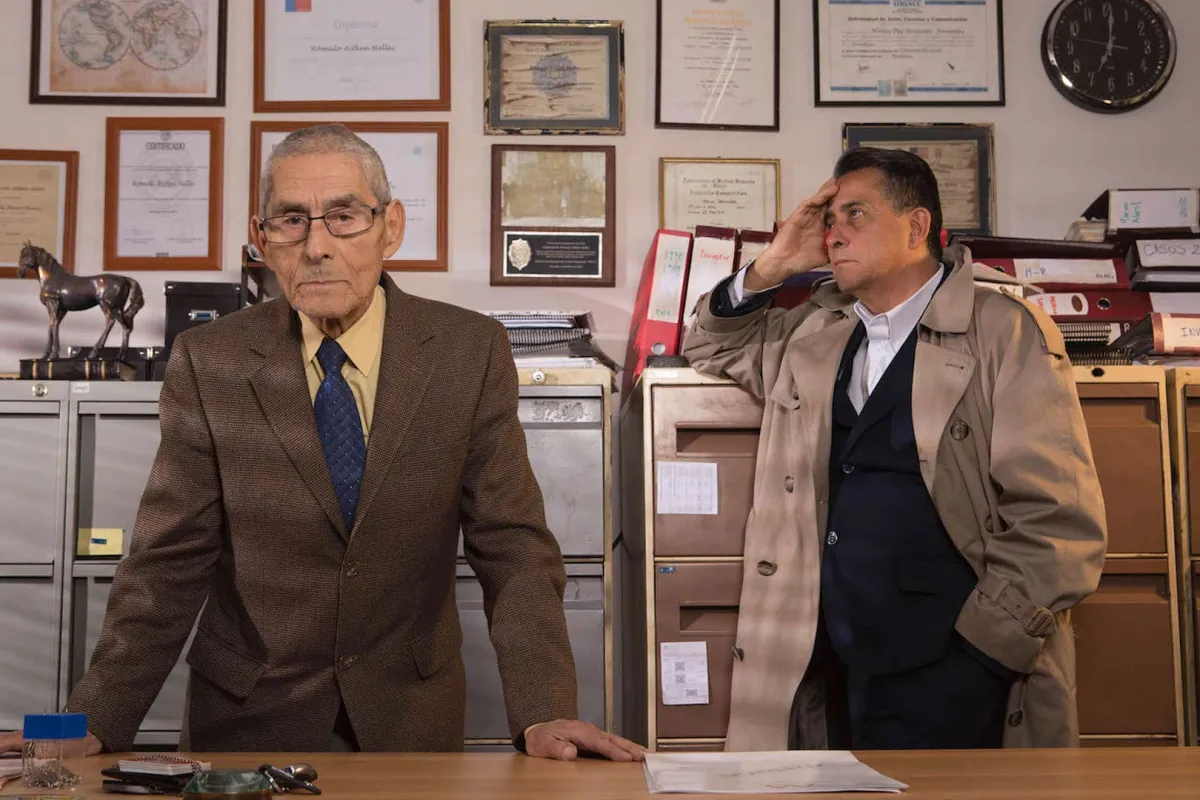 An elderly man in a brown suit and a middle-aged man in trench coat are behind an office desk