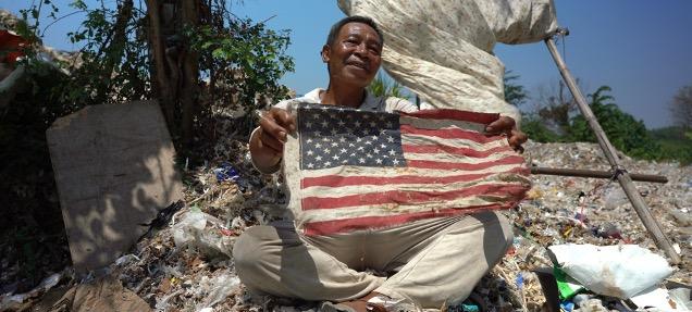 An Indonesian man holds a tattered American flag in a trash heap. 