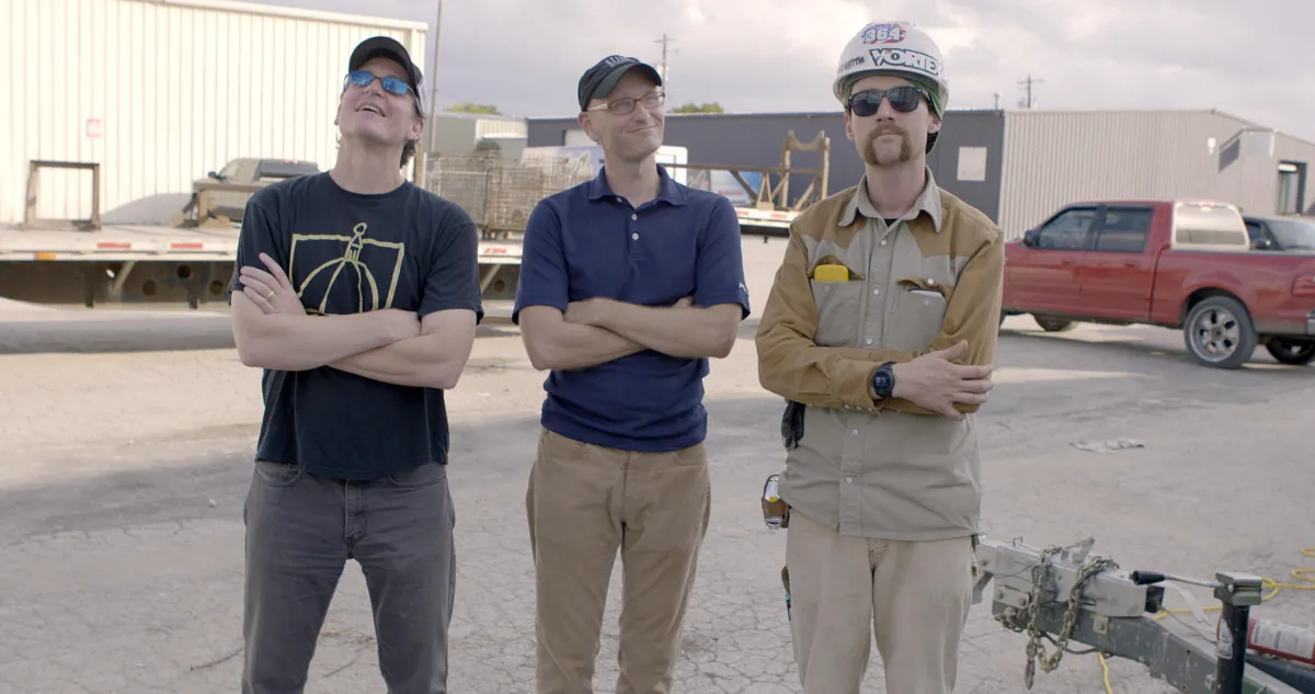 Three Caucasian male are smiling at the camera with their arms interlocked.
