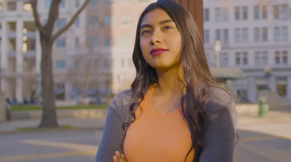 A young Latina woman stands with arms crossed in front of distant buildings