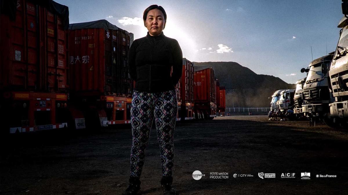 woman stands in front of trucks, mountains in the background