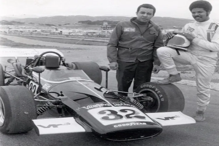 two black male racers stand next to a racecar at a speedway.
