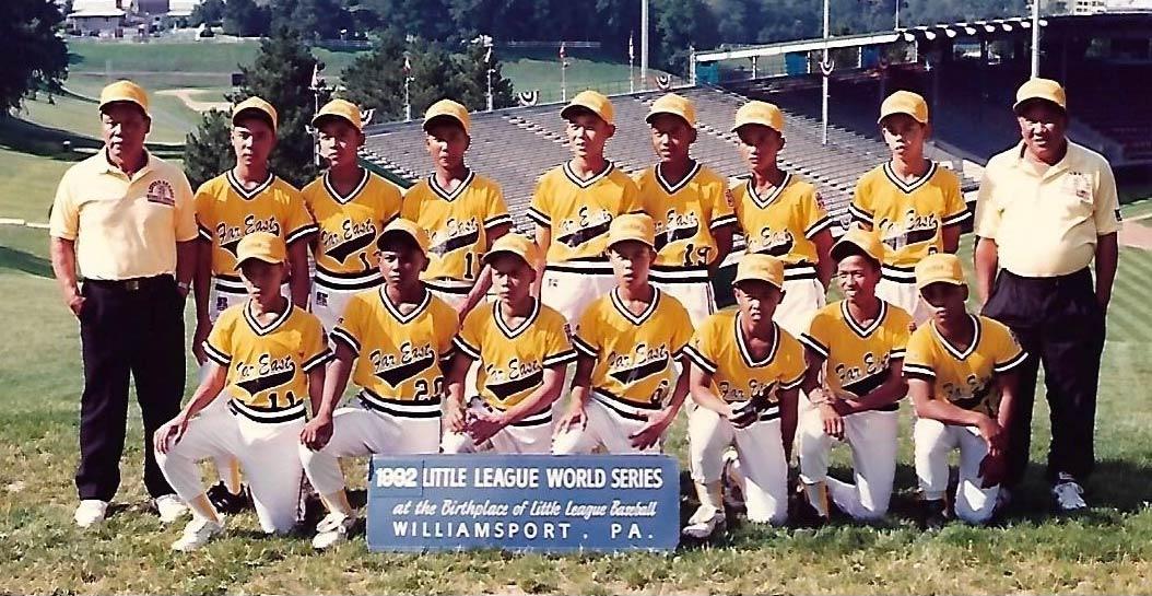 The 1992 Little League World Series Champion team from the Philippines pose in front of the baseball stadium in yellow jerseys and hats. Their coaches stand on either side of the players. 