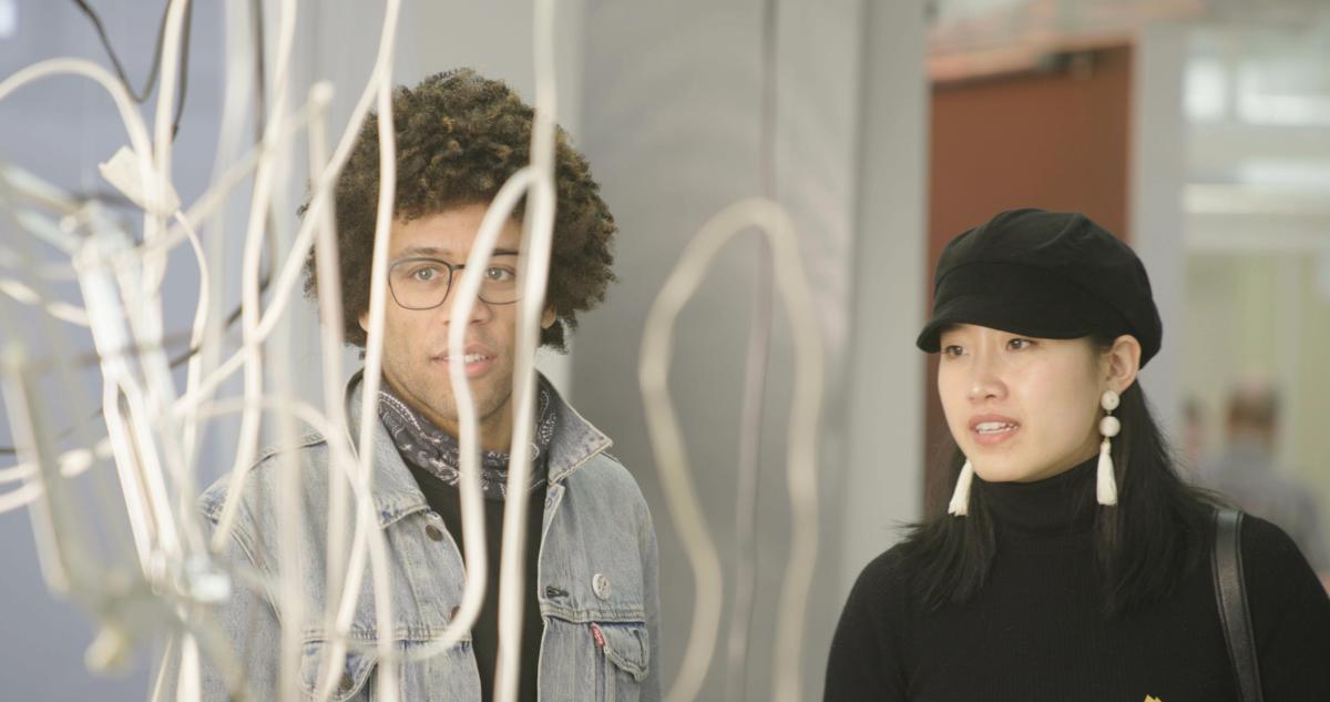 A young Black man with curly hair, glasses and a scarf and a young Asian woman with a black cap and white earrings stand before an abstract white curly art piece in a museum.