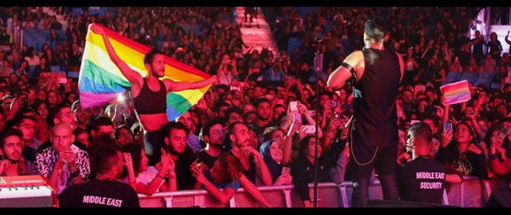 A singer in a leather outfit stands before a crowd of fans, one waving a Pride Flag over their shoulders.