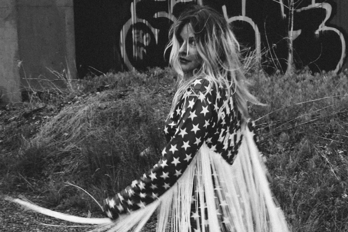 Elizabeth Cook twirls in front of a graffitied wall wearing a star studded jumpsuit with tassels