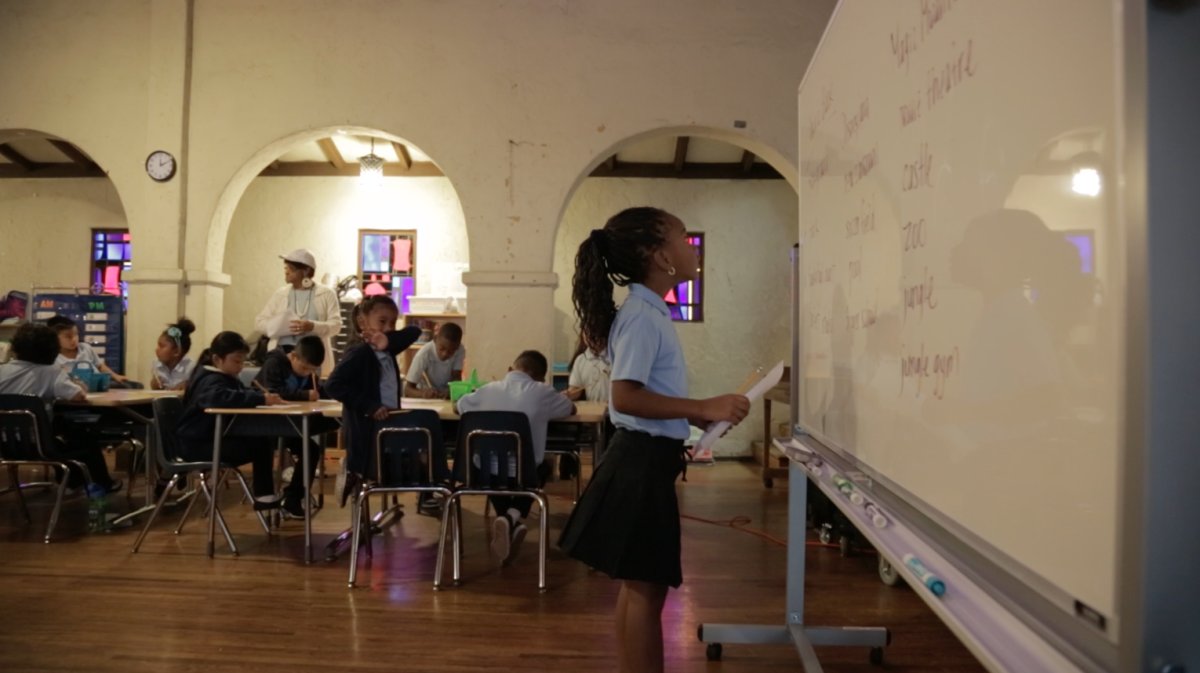 a grade school African American girl in a school uniform looks up at a white board with a paper in her hand in front of the rest of her young classmates.
