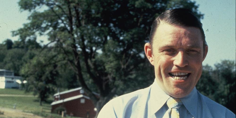 A Middle-aged White man smiles in a blue button-up and yellow tie in front of a rural set of houses.