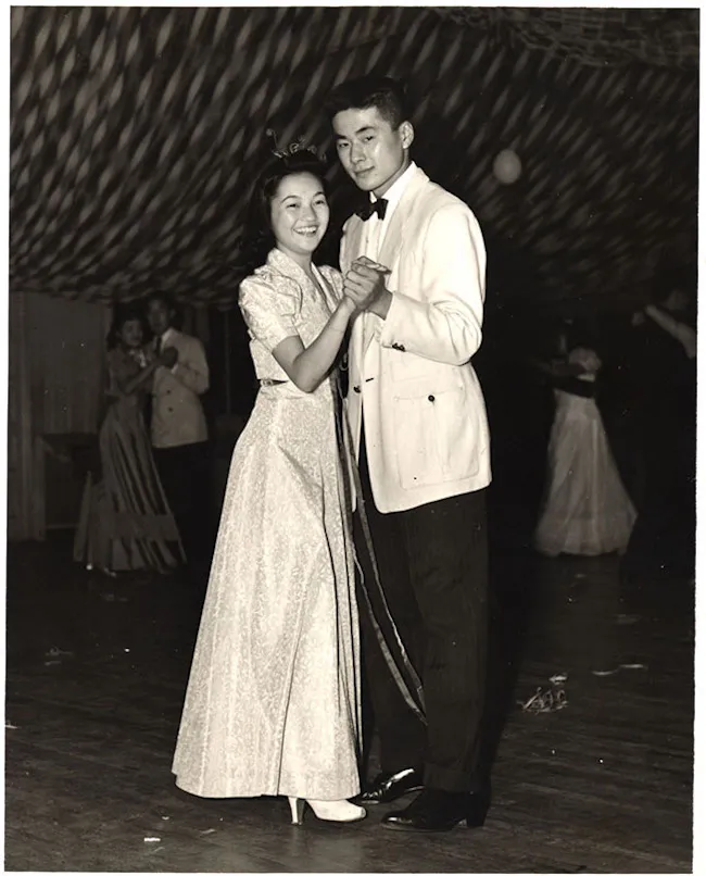 Mary and Kelly Adachi smile and slow dance in their wedding outfits. 