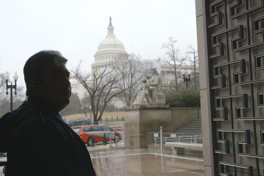 A man is in the shadows standing at the doorway with the US State Capital across the street
