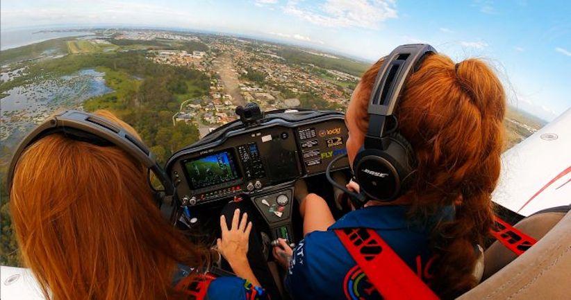 two red-headed women at the controls of an aircraft in midflight - green landscape stretches out beyond the clear glass cockpit window.