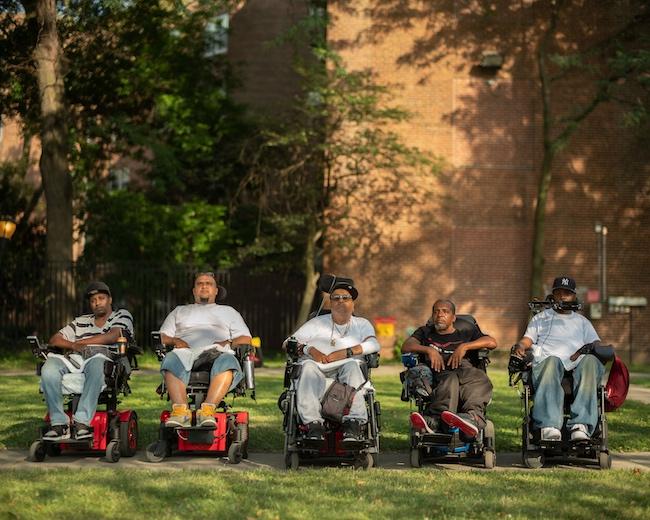 Five Black and brown men (from left to right: Vince, Jay, Tito, Pete and Var) sit in wheelchairs outside, lined up in a straight horizontal line facing the camera. They wear T-shirts and sneakers and some wear baseball caps. They all look into the camera with serious expressions. 