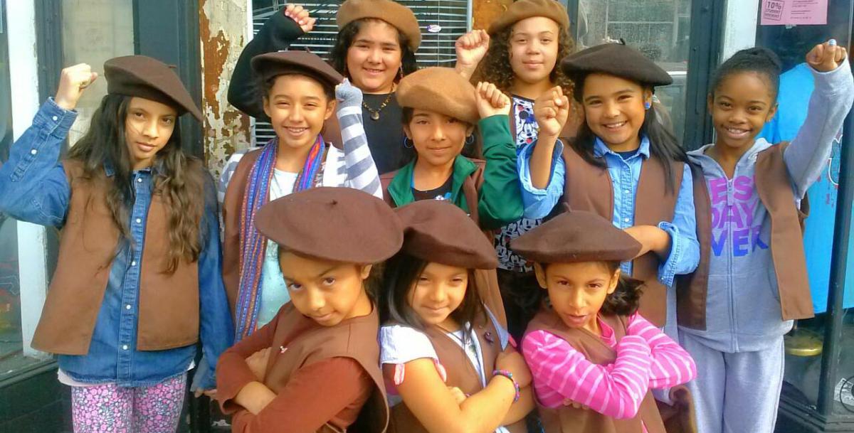 A group of young girls of color stand in brown berets with their fists raised. 