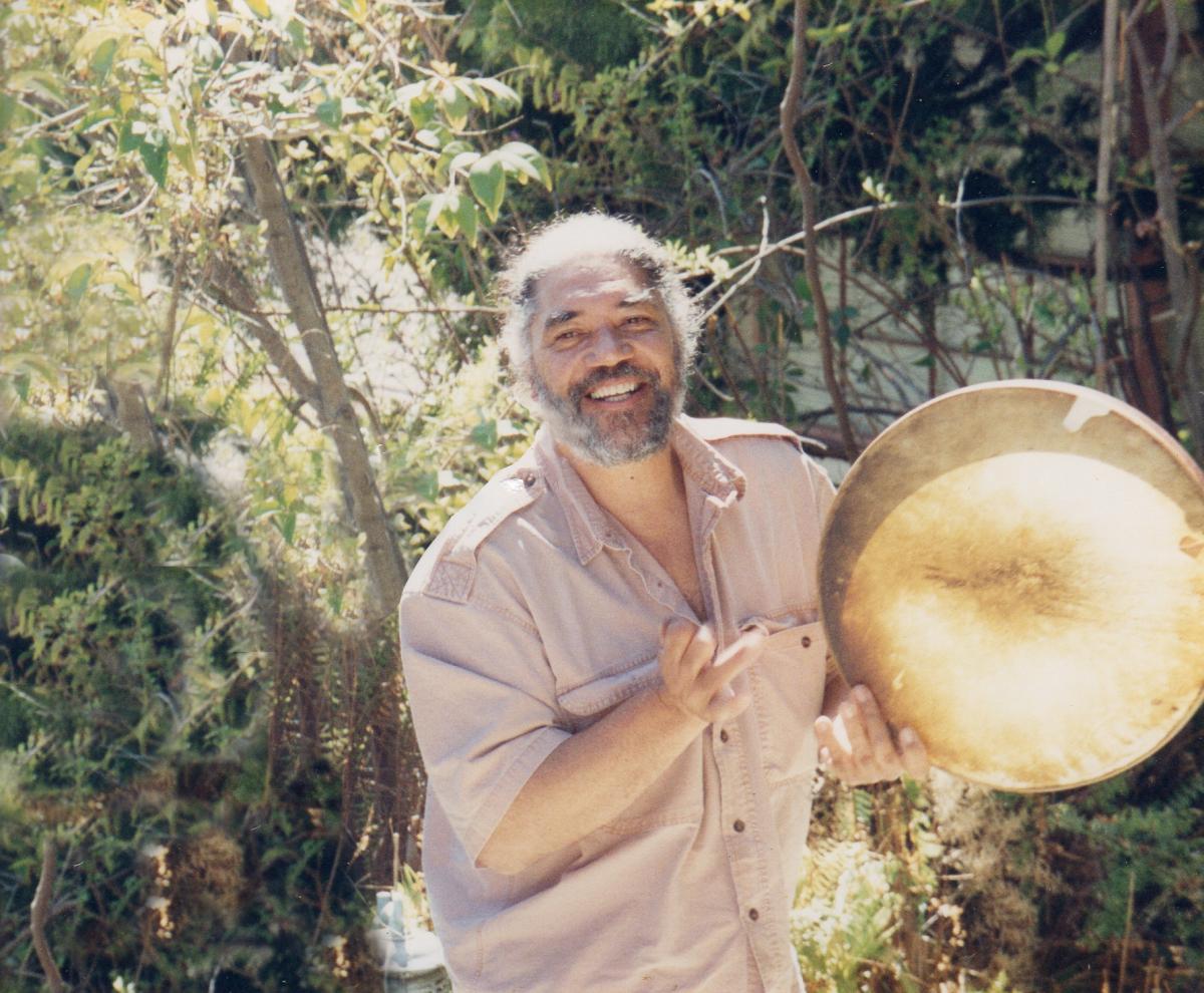 Bruce stands holding a drum before a forest.