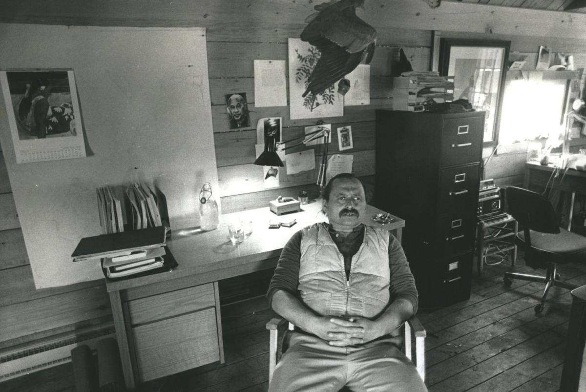 Jim Harrison sits with his hands in his lap in his rugged, outdoorsy 20th-century office. 