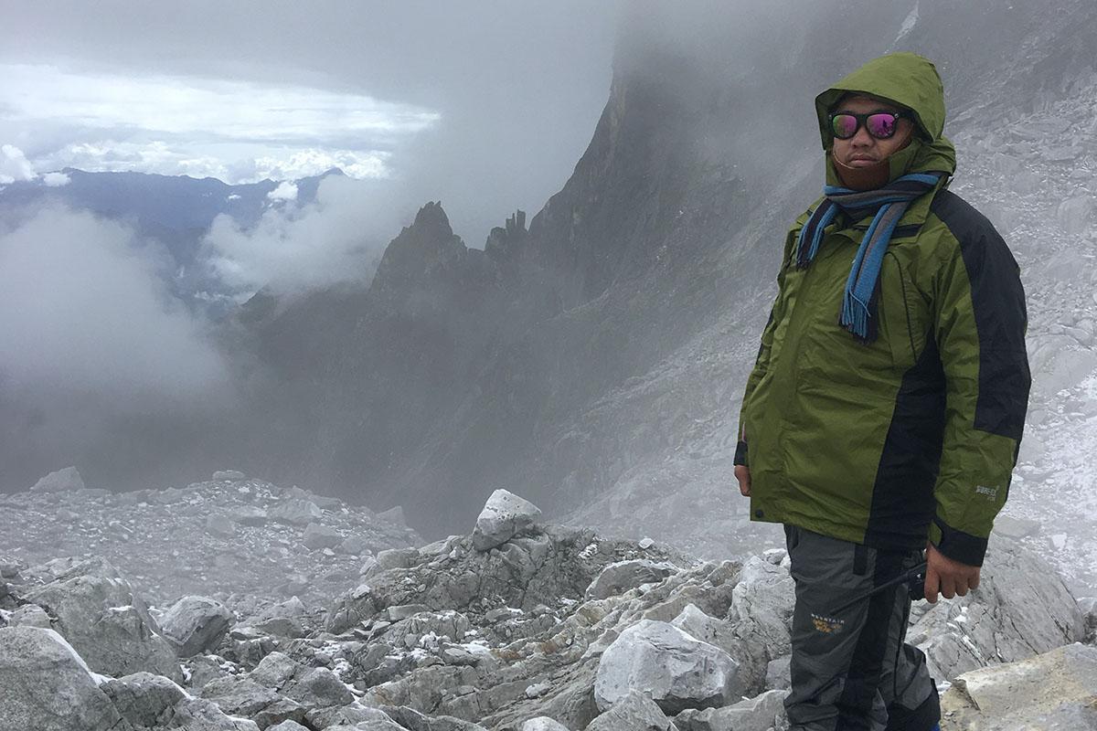 Phuntsho Tshering, a man of South Asian descent, is standing on top of a rocky terrain in the rugged mountainous Himalayas looking straight into the camera. He is wearing shades, a pair of gumboots and hiking clothes. 