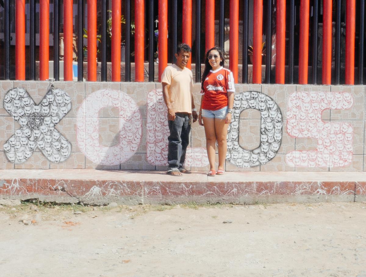 Two people are standing in front of a wall that has 'xolos' graffitied on it.